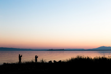 Silhouettes of a couple on a lake shore taking photos of sunset 