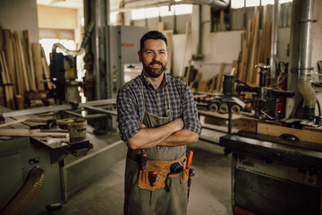 Portrait of a smiling young woodworker standing with his arms crossed by a bench saw in his...