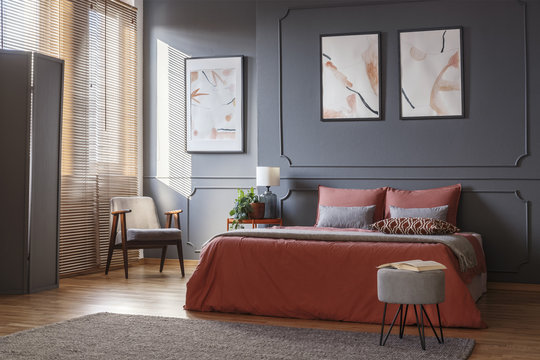 Gray, retro armchair standing in the corner of an elegant bedroom interior with watercolor posters on dark gray wall with molding and orange sheets on the bed. Real photo
