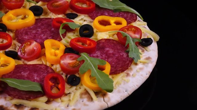 Closeup view of tasty homemade pizza with salami and vegetables rotating on black background in 4K. Traditional tasty food.
