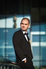 handsome stylish groom sitting on modern metal handrails holding hands in pockets of elegant suit