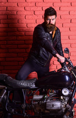 Masculine passion concept. Hipster, brutal biker on serious face in leather jacket gets on motorcycle. Man with beard, biker in leather jacket near motor bike in garage, brick wall background.