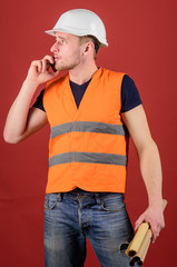 Man, foreman in helmet speaking on phone, red background. Architect calling engineer to fix plan. Engineer, architect, on busy face speaks on smartphone while holds blueprints. Communication concept.