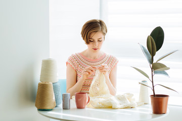 Сraftswoman knitting dress with crochet in cozy workplace at home interior. Female working with tender lace. Business handmade crochet relaxation concept.