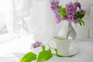 white cup with tea and a bouquet of lilac in a white vase on a summer morning breakfast at the window with a white lacy curtain in a rustic style with a copy of the space
