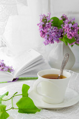 white cup with tea and a bouquet of lilac in a white vase on a summer morning breakfast at the window with a white lacy curtain in a rustic style with a copy of the space