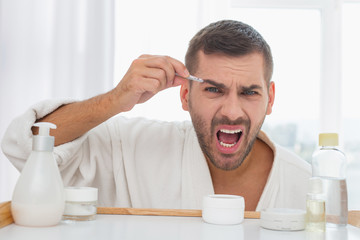 Handsome look. Joyful nice man smiling while plucking eyebrows with his tweezers