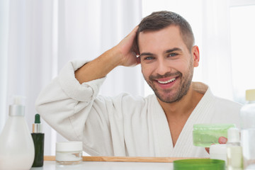 Stylish look. Positive handsome man holding a bottle of hair gel while standing in front of the mirror