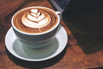 Closeup image of a cup of hot latte coffee with heart latte art  and laptop on wooden table