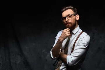 Fotobehang Young stylish hipster with cool hairstyle and beard dressed in white shirt and suspenders is thinking of a new creative idea looking at viewer © berezko