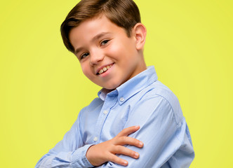 Handsome toddler child with green eyes with crossed arms confident and happy with a big natural smile laughing over yellow background