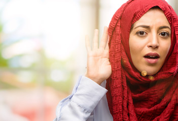 Young arab woman wearing hijab holding hand near ear trying to listen to interesting news...