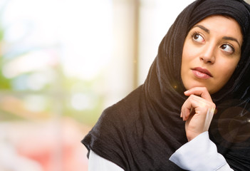 Young arab woman wearing hijab thinking and looking up expressing doubt and wonder