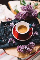Fototapeta na wymiar Delicious fresh morning coffee in a ceramic cup with a beautiful crema, reading glasses and a purple blossoming lilac on the black plaid kitchen cloth on the wooden slab table background 