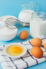 Eggs of chicken, flour in a bowl, sugar, milk in a glass jar whisk ingredient for cooking a bake on a blue background with copy space