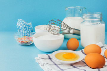 Eggs of chicken, flour in a bowl, sugar, milk in a glass jar whisk ingredient for cooking a bake on a blue background with copy space