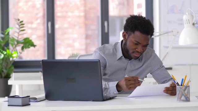 business, people, paperwork and technology concept - african american businessman with laptop computer and papers working at office