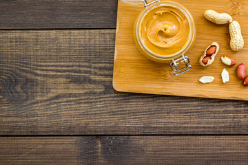 Make peanut butter. Paste in glass jar on cutting desk near peanut on dark woden background top view copy space