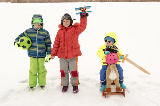 Funny girl in flying glasses and helmet pilot. Boy is sitting on oy horse with sword. The girl plays with plane as pilot, and child presents herself as knight. Brother is standing with green ball.