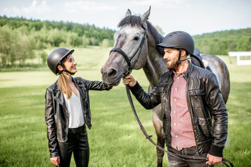 Young couple riders standing near the beautiful horse on the green meadow