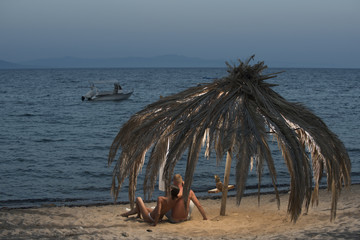Fototapeta na wymiar Topless woman and man lie under umbrella made out of dried palm leaves. Love concept. Couple in love full of desire at beach, sea background. Couple on vacation at tropical seashore, honeymoon.