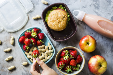 Cooking lunch for the child to school. On the gray kitchen table. Sandwich, strawberries and peanuts in lunchboxes. Top view. Woman puts strawberries in a container