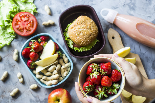 Cooking lunch for the child to school. On the gray kitchen table. Sandwich, strawberries and peanuts in lunchboxes. Top view. Woman holding a bowl of strawberries