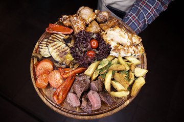 A variety of baked meat, fish, vegetables on a wooden board in the hands of the waiter.