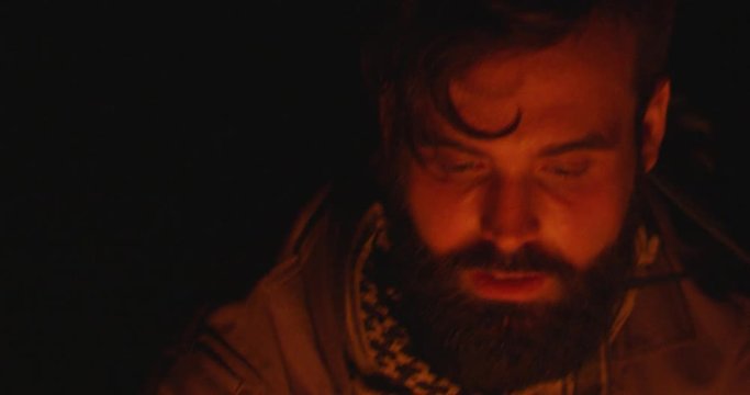 Bearded man sits near campfire on a campaign and lights a bonfire at night