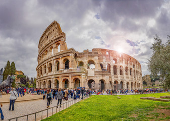 tourists walking under the colosseum in rome on a cloudy day with the sun shining behind