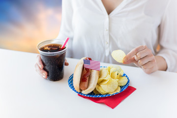 fast food, unhealthy eating and people concept - close up of woman with hot dog, cola in plastic cup and chips on independence day over evening sky background