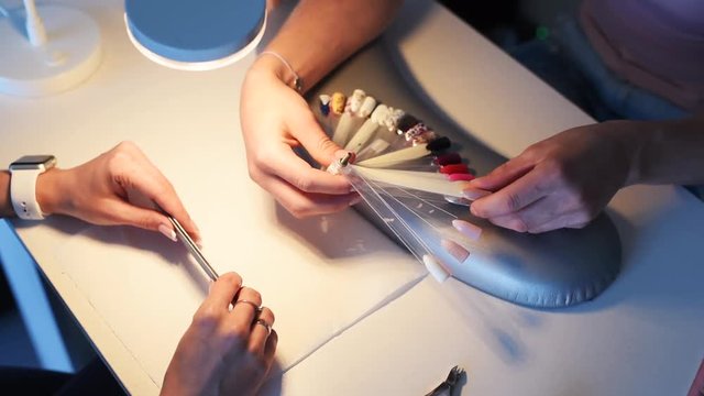 Close-up of female client's hands choosing color and design from artificial nails palette samples for gel manicure in a beauty salon. Professional advises what color to choose.