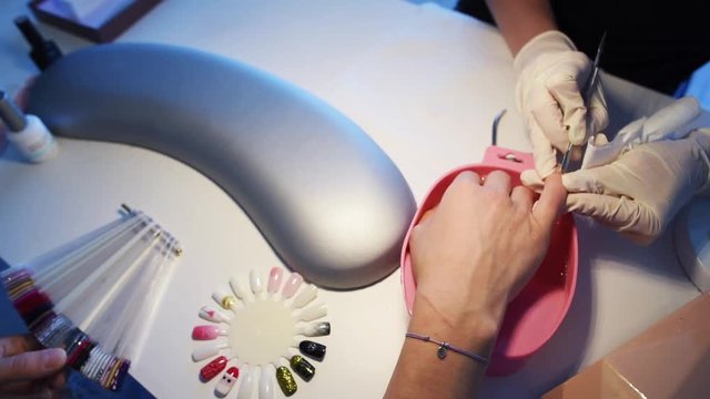 Manicurist in disposable gloves using cuticle pusher for pushing cuticles on client’s little finger while customer is choosing gel polish color from sample palette. Slow motion.