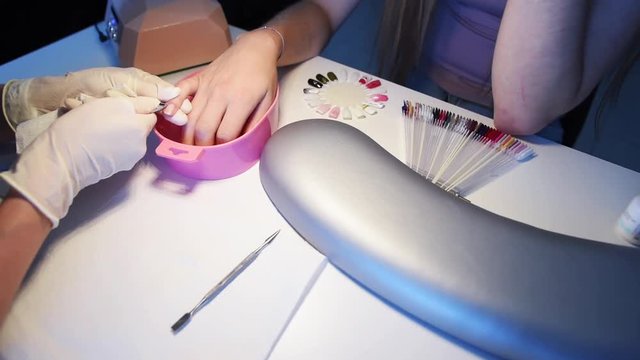 Close-up of nail master hands in disposable gloves cutting cuticles by using manicure cutters on client’s fingers while she is choosing gel polish color from sample palette.