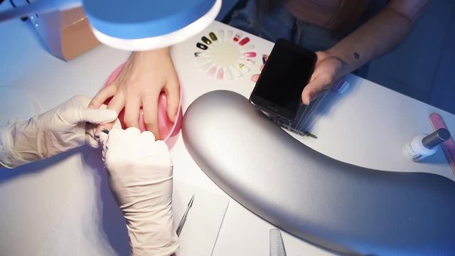 Close-up of nail master hands in disposable gloves cutting cuticles by using manicure cutters on client’s fingers while customer is looking at cell phone. Slow motion
