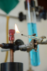 Flame burning from a glass tube with laboratory equipment as a diffuse background.