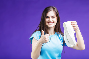 Pretty happy smiling brunette girl dressed in blue t-shirt holding sport footwear sneakers before blue background