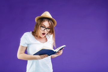 Pretty blonde girl dressed in white t-shirt, hat and eyeglasses reads book with excited face expression before blue background