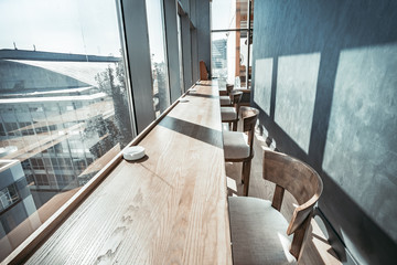 Close-up view of the contemporary restaurant interior the long wooden table with the comfortable chairs. Wonderful panoramic view from the window. Stylish decor in grey shades.