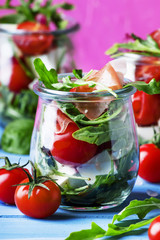 Salad with arugula, cheese, cherry tomatoes and prosciutto in glass jars, pink blue background, selective focus