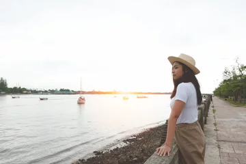 Foto op Canvas Side view of happy asian woman in hat standing and looking at river in evening sunset. © AePatt Journey