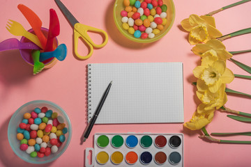 The concept of children's creativity. On a pink background color paints, scissors, in multi-colored plates a colorful color dragee, flowers narcissuses, a notebook with a pen.