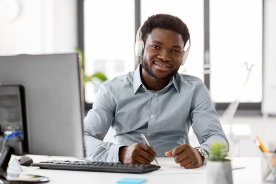 business, technology and people concept - happy african american businessman with headphones and papers listening to music at office