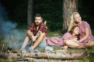 Bearded man cooking sausages on sticks above fire while his female friends are having rest. Brunette girl with curly hair lying next to her blond smiling friend