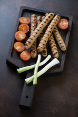 Roasted pork sausages with cherry tomatoes and onion on a wooden serving tray, vertical shot on a brown metal background