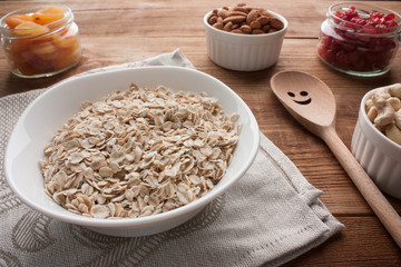 Oat flakes in bowl with dried apricots, dried cherries, almonds, cashews in jars on wooden table with wooden spoon.