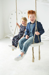 Cute kids red boy brother and blonde sister have a fun on the swing in the white studio