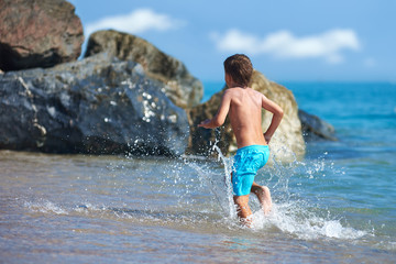 Cute Caucasian boy is running in the water along the sea shore against big boulders.
