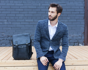 A young businessman with a beard in a fashionable suit is resting sitting on a wooden bench in the business center of the city.