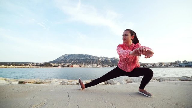 Young fit woman doing stretching exercises for legs after running by the sea with mountain on background.